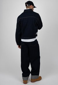 Homme portant une veste en denim foncé et un pantalon avec les revers roulés, des bottes couleur sable, une casquette noire et une chemise blanche, debout dos à la caméra sur fond gris.