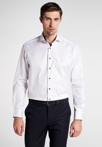 Weißes Langarmhemd mit schwarzen Knöpfen, Button-Down-Kragen und glattem Stofffinish, kombiniert mit dunklen Hosen.
