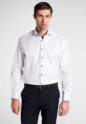 Weißes Langarmhemd mit schwarzen Knöpfen, Button-Down-Kragen und glattem Stofffinish, kombiniert mit dunklen Hosen.