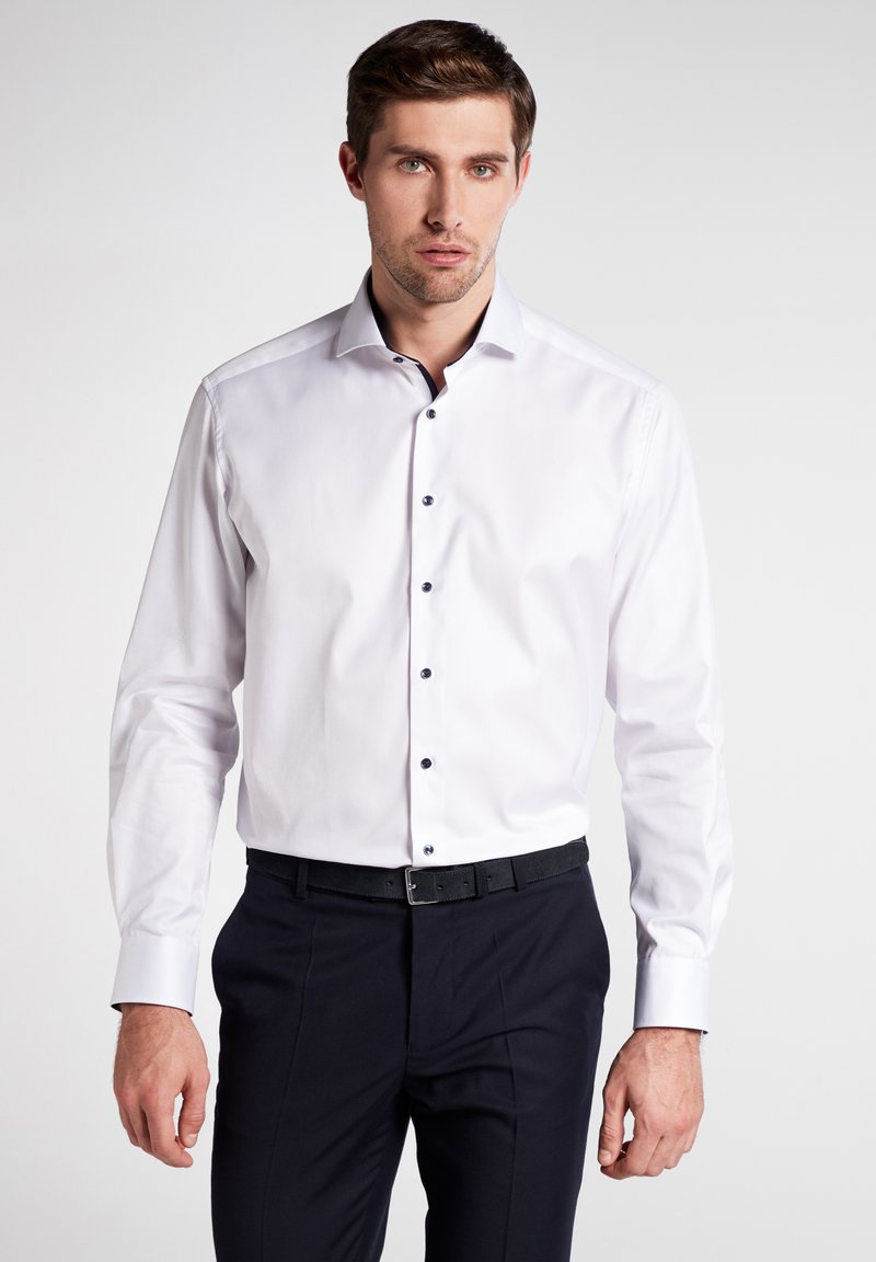 Weißes Langarmhemd mit schwarzen Knöpfen, Button-Down-Kragen und glattem Stofffinish, kombiniert mit dunklen Hosen.