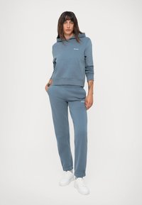 Světle modrý dvoudílný outfit zahrnující mikinu s kapucí a přední kapsou, k tomu přizpůsobené joggingové kalhoty s elastickým pasem, doplněné o bílé tenisky.