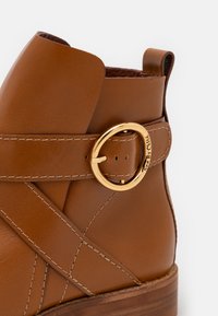 Bottine en cuir marron avec des brides cousues, une boucle circulaire dorée gravée "See By Chloé" et un talon empilé en bois.