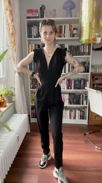 Sort jumpsuit med korte, voluminøse ærmer og dyb v-udskæring. Bæres med flerfarvede sneakers. Baggrunden viser bogreoler og stueplanter.