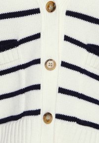 Bílý pletený cardigan s horizontálními námořnickými pruhy a třemi hnědými knoflíky se čtyřmi otvory uprostřed vpředu.