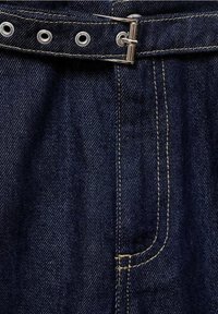 Tejido de mezclilla oscura con textura de tejido visible, que presenta herrajes plateados, un lazo para cinturón con ojales y costuras amarillas en contraste.