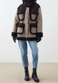 Persona vestida con un abrigo de cuadros negro y beige con ribetes negros, jeans azules, guantes negros y botas de cordones negras, de pie sobre un suelo beige.