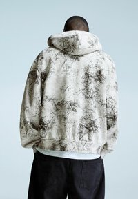 Kaptrui in lichtbeige met donkere spetterpatronen; casual pasvorm, geribbelde boorden en onderrand; gedragen met zwarte broek; gestructureerde stof.