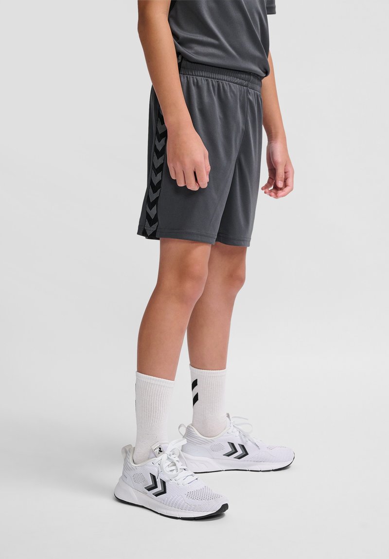 Grijze sportshorts met zwarte chevronaccenten, gecombineerd met witte sneakers en witte sokken met zwarte strepen. Casual, aansluitend ontwerp.