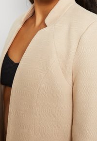 Chaqueta texturizada beige con un patrón de espiga, que presenta un cuello chal y un diseño ajustado, parcialmente mostrada contra una parte superior oscura.