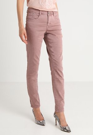 Kobieta ubrana w obcisłe, różowe dżinsy w kolorze pyłowego różu oraz wysokie buty na obcasie w wężowy wzór, stojąca na jednolitym białym tle.