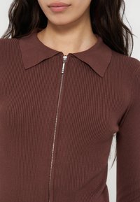 Femme portant un haut marron côtelé zippé avec col, montrant le haut du torse et une partie du visage avec de longs cheveux foncés.