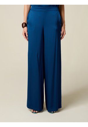Femme portant un pantalon bleu à jambes larges et des sandales bleues, debout les mains relâchées le long du corps sur un fond uni.