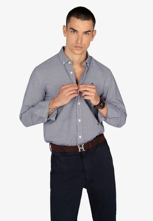 Camisa de cuadros en azul marino y blanco con botones, mangas largas, cuello ligeramente marcado y corte ajustado. Combinada con pantalones oscuros y un cinturón marrón con hebilla de plata.