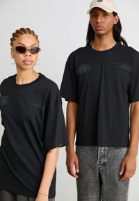 Tricouri negre oversized din bumbac, cu un logo și text imprimate pe partea din față. Modelul poartă ochelari de soare; fundalul este simplu.