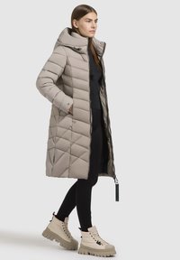 Dolga, svetlo bež pufferska bunda s kvadratnim vzorcem, prevelikim kapucinom in zadrgo spredaj. Model nosi črne legice in debele kremne škornje.