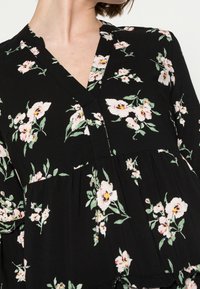 Blouse noire avec un col en V, présentant un motif floral avec des accents roses et verts, des manches évasées et un ourlet froncé. Texture de tissu douce.
