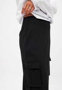 Svarta byxor med en slät yta, sidofickor med klaffar och en figurnära design. Midjebandet sitter i det naturliga midjebandet.