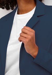 Granatowa marynarka z klapkami, zapinana na jeden guzik, z gładkiego materiału, nałożona na białą koszulkę, ręka trzymająca kołnierz.