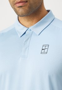 Lyseblå polo skjorte med et strukturert mønster, knapper ved kragen, og en liten svart Nike-logo på venstre brystområde.