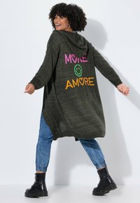 Žena se usmívá a obrací se, na sobě má zelený kardigan s kapucí, na kterém je nápis "MORE" a "AMORE" spolu s úsměvovým obličejem, modré džíny a černé boty.