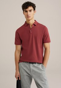 Camiseta polo burdeos con cuello y tapeta de tres botones; mangas cortas; combinada con pantalones gris claro; textura suave.
