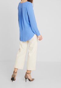 Světle modrá halenka s dlouhými rukávy a zaobleným lemem, v kombinaci se smetanovými širokými kalhotami a vysokými podpatky se vzorem hadí kůže. Hladká textura.