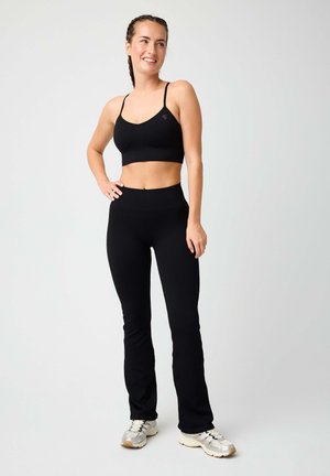 Frau lächelt, trägt ein schwarzes, figurbetontes Crop-Top mit Spaghettiträgern, passende schwarze Hose und weiße Sneakers, steht vor einem schlichten Hintergrund.