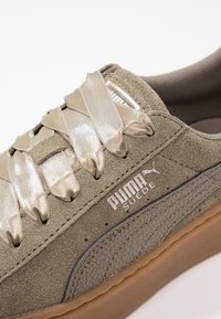 Semišové tenisky v odstínu tan s metalickými saténovými tkaničkami, texturovanou gumovou podrážkou a bílým logem na boku. Obsahují prošívané detaily.