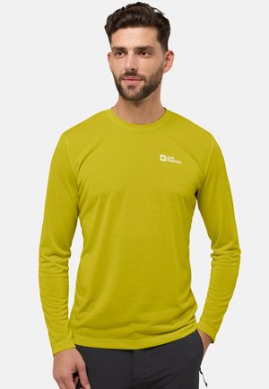 Man met baard in een limoen groene longsleeve met het logo 'Jack Wolfskin', staand met één hand in de zak van zijn donkere broek tegen een witte achtergrond.