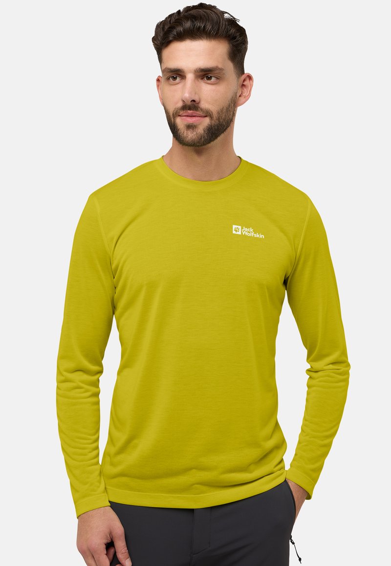 Man met baard in een limoen groene longsleeve met het logo 'Jack Wolfskin', staand met één hand in de zak van zijn donkere broek tegen een witte achtergrond.
