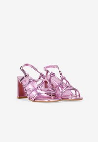Sandali con tacco metallici rosa, con design a laccetto e dettagli annodati, dotati di cinturino regolabile alla caviglia. Finitura liscia e tacco a blocco.