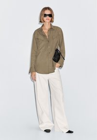 Olive green button-up shirt with two chest pockets, white wide-leg trousers, black handbag, and sleek black sunglasses. Smooth fabric texture.