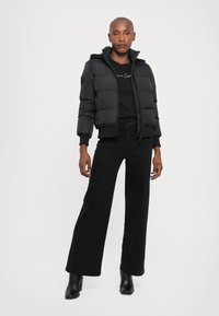Doudoune noire avec capuche, fermeture éclair à l'avant et poignets côtelés. Portée par-dessus un haut noir et associée à un pantalon large noir et des bottes à talons.