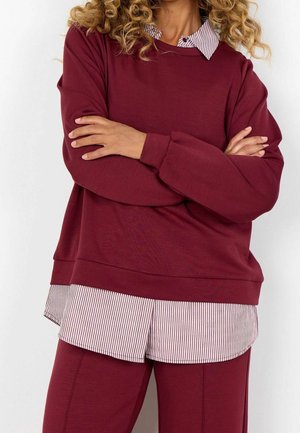 Person with curly hair wearing a burgundy sweatshirt over a striped shirt, with arms crossed, shown from neck to mid-thigh.