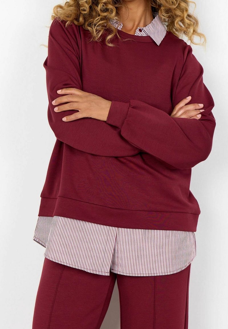 Personne aux cheveux bouclés portant un sweat-shirt bordeaux par-dessus une chemise rayée, les bras croisés, vue du cou jusqu'au milieu des cuisses.