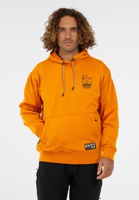 Mann mit lockigem Haar, der einen orangefarbenen Kapuzenpullover mit Brustdruck und schwarze Hosen trägt, vor einem weißen Hintergrund stehend.