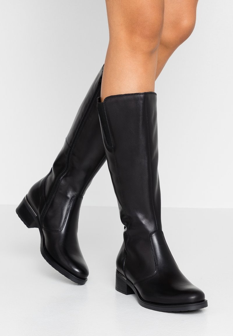 Mustat nahkaiset polvipituiset saappaat, joissa on matalat korot, päällä henkilö, joka seisoo valkoisella lattialla harmaata taustaa vasten.