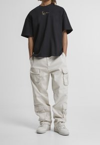 Persona con camiseta negra de gran tamaño con logo en cursiva, pantalones cargo beige con múltiples bolsillos y zapatillas blancas, con las manos en los bolsillos.