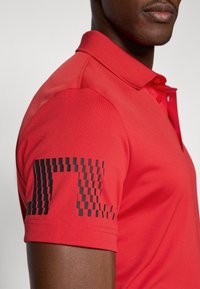 Camisola polo vermelha com uma pala de 3 botões e mangas curtas. Apresenta um padrão geométrico preto na manga esquerda, com uma textura de tecido suave.