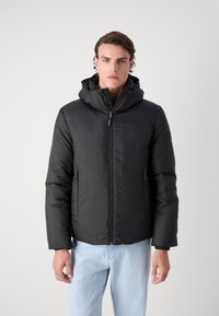 Svart boblejakke med hette, med glidelås foran, sidelommer og en myk, glatt tekstur. Bært med lyseblå jeans.