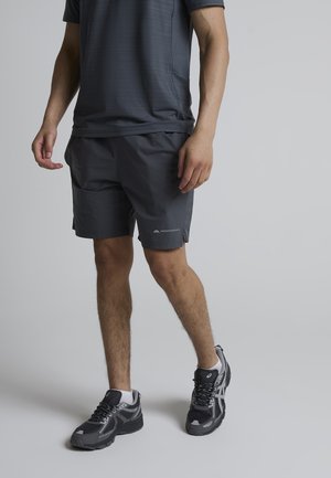 Mand iført mørkegrå atletiske shorts og matchende t-shirt med sorte og grå løbesko, stående på en lysegrå gulv.