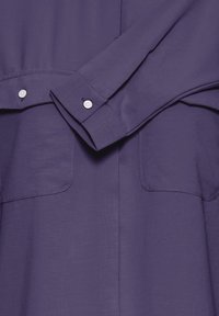 Camicia a maniche lunghe viola scuro con bottoni bianchi sui polsini e due tasche a toppa anteriori, texture del tessuto visibile.