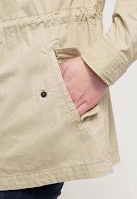 Beige lättviktsjacka med dragsko i midjan och sidoficka. Har en texturerad yta och en knappdetalj nära fickan.