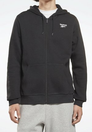 Mann trägt schwarzen Reebok Zip-Hoodie mit Fronttaschen und hellgraue Jogginghose, stehend vor schlichtem Hintergrund.