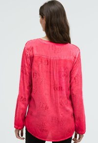 Frau mit langen braunen Haaren, die eine lockere, langärmlige, leuchtend pinke Bluse mit gedruckten Wörtern wie "Liebe", "Glücklich" und "Je t'aime" trägt, von hinten gesehen.