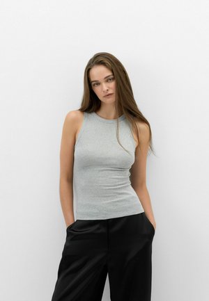 Giovane donna con lunghi capelli castani che indossa un top grigio senza maniche e pantaloni neri in piedi contro un muro bianco semplice.
