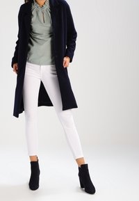 Navy long coat, light green gathered neck top, white slim-fit jeans, and black ankle boots with a block heel. Clean design and tailored fit.