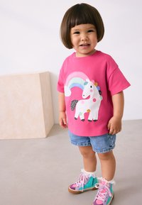 T-shirt en coton rose avec un motif licorne et arc-en-ciel, associé à un short en denim et des baskets montantes colorées avec des accents en dentelle.