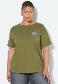 Osoba s krátkými vlasy nosí olivově zelené tričko s fialovým designem oka na hrudi, tmavé džíny, zlaté kruhové náušnice a prsteny na prstech.