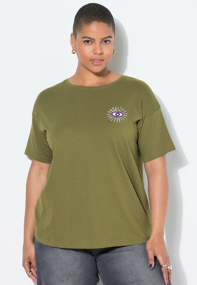 Osoba s krátkými vlasy nosí olivově zelené tričko s fialovým designem oka na hrudi, tmavé džíny, zlaté kruhové náušnice a prsteny na prstech.